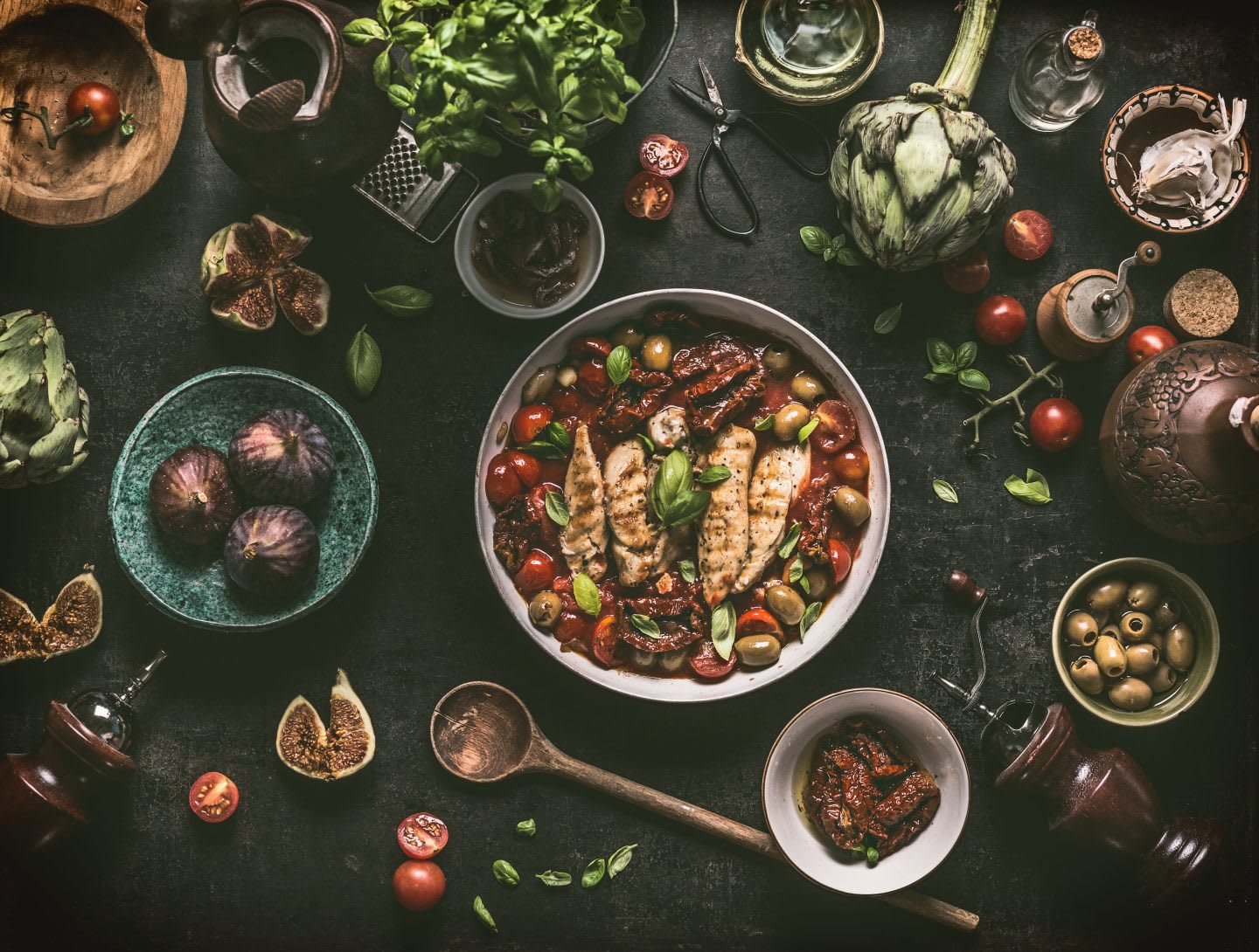 Grilled chicken breast with Mediterranean sauce in cooking pan and ingredients: tomatoes, artichokes, olives, sun-dried tomatoes,figs, anchovies, fresh seasoning herbs and spices on dark rustic table with vintage kitchen utensils , top view.