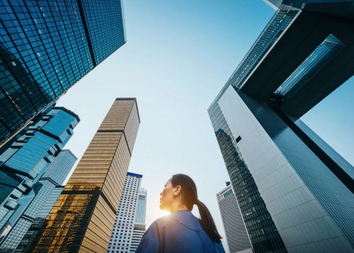 Low angle view of woman standing against modern skyscrapers and blue sky in city at sunrise. relocation, city, confidence, international, working abroad, GamePlan A