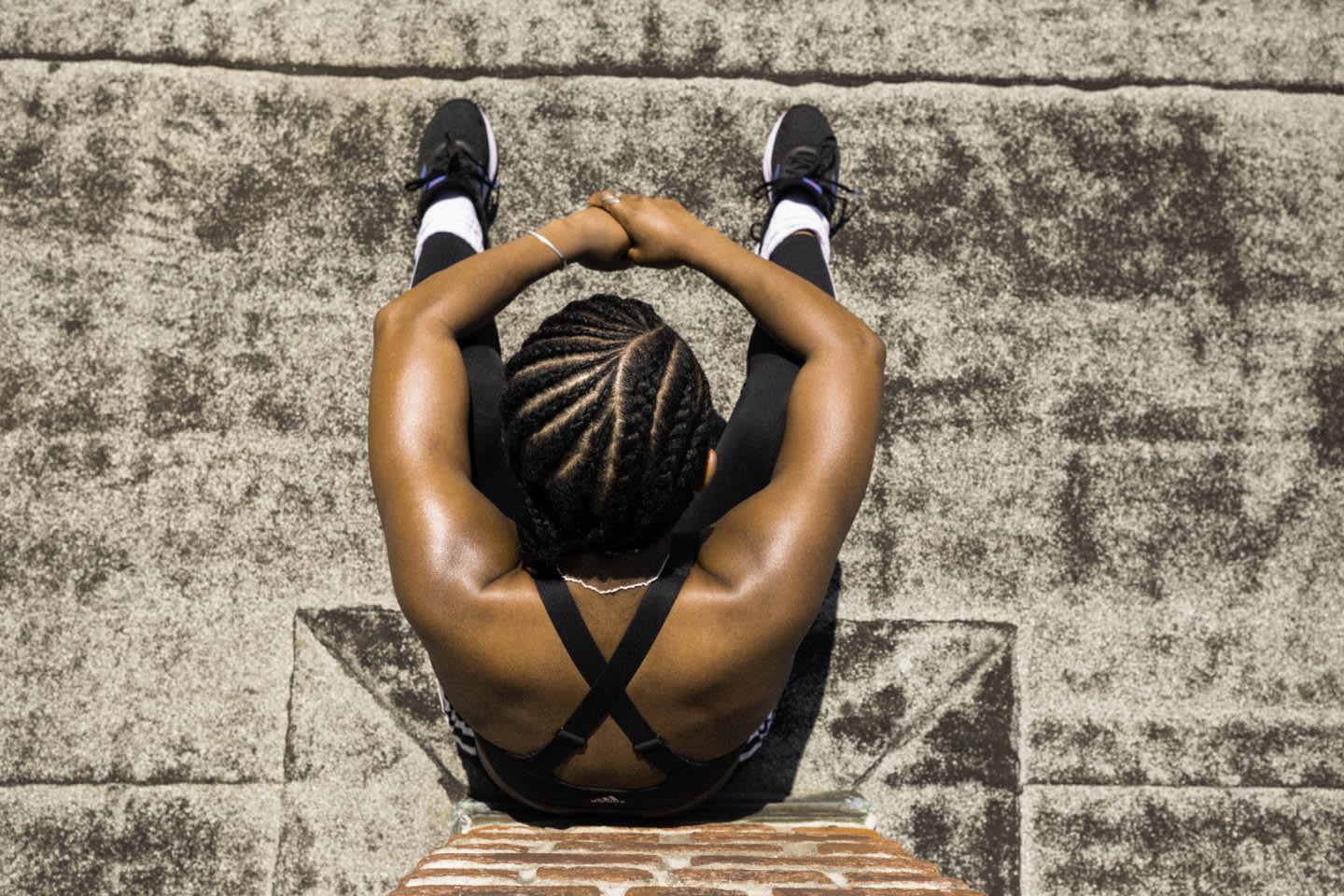 Woman wearing black sports bra, black running tights and black running shoes sitting with knees up against brick wall in the sun, athlete, rest, recovery, tired, adidas, sport, fitness