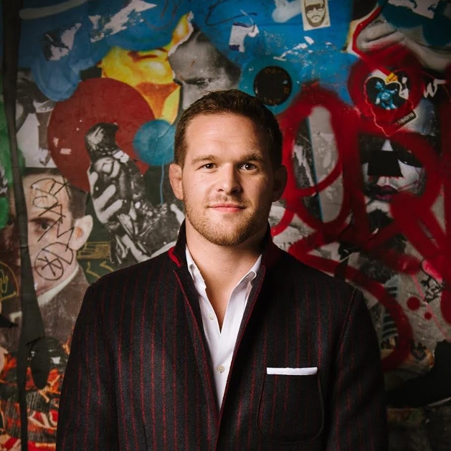 Headshot of Caucasian man wearing dark suit standing in front of graffiti wall, Hudson Taylor, founder, Athlete Ally, LGBTQ, charity