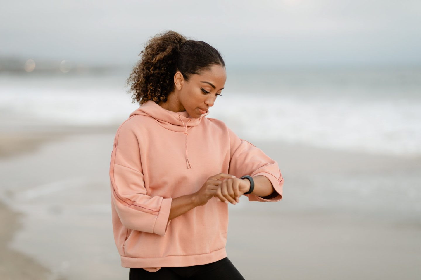 Woman wearing light pink hoodie checking her watch at the beach, outdoors, beach, seaside, woman, girl