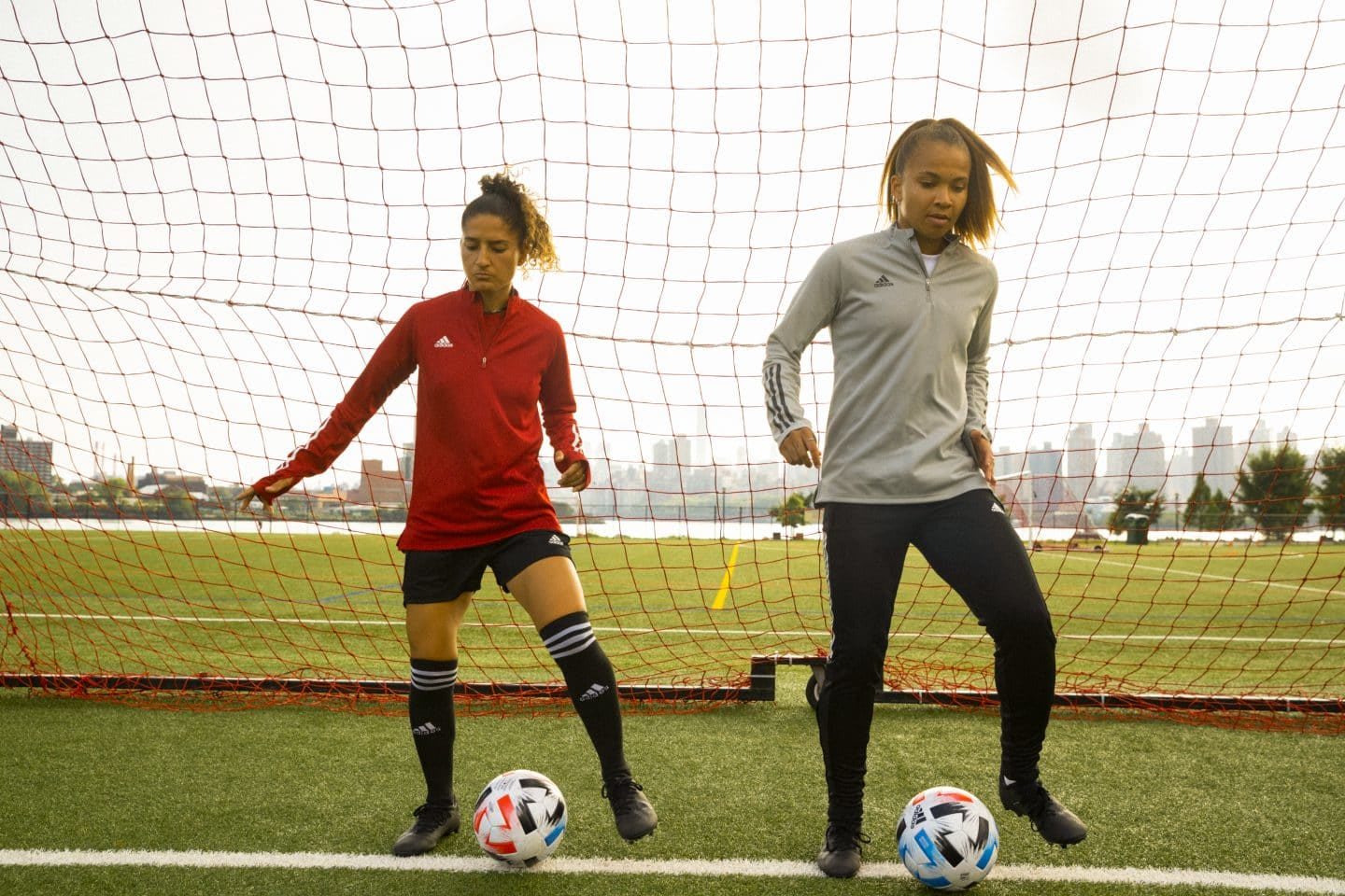 Two women dribbling footballs on a field in front of a goal, sports, football, soccer, exercise, fitness, adidas