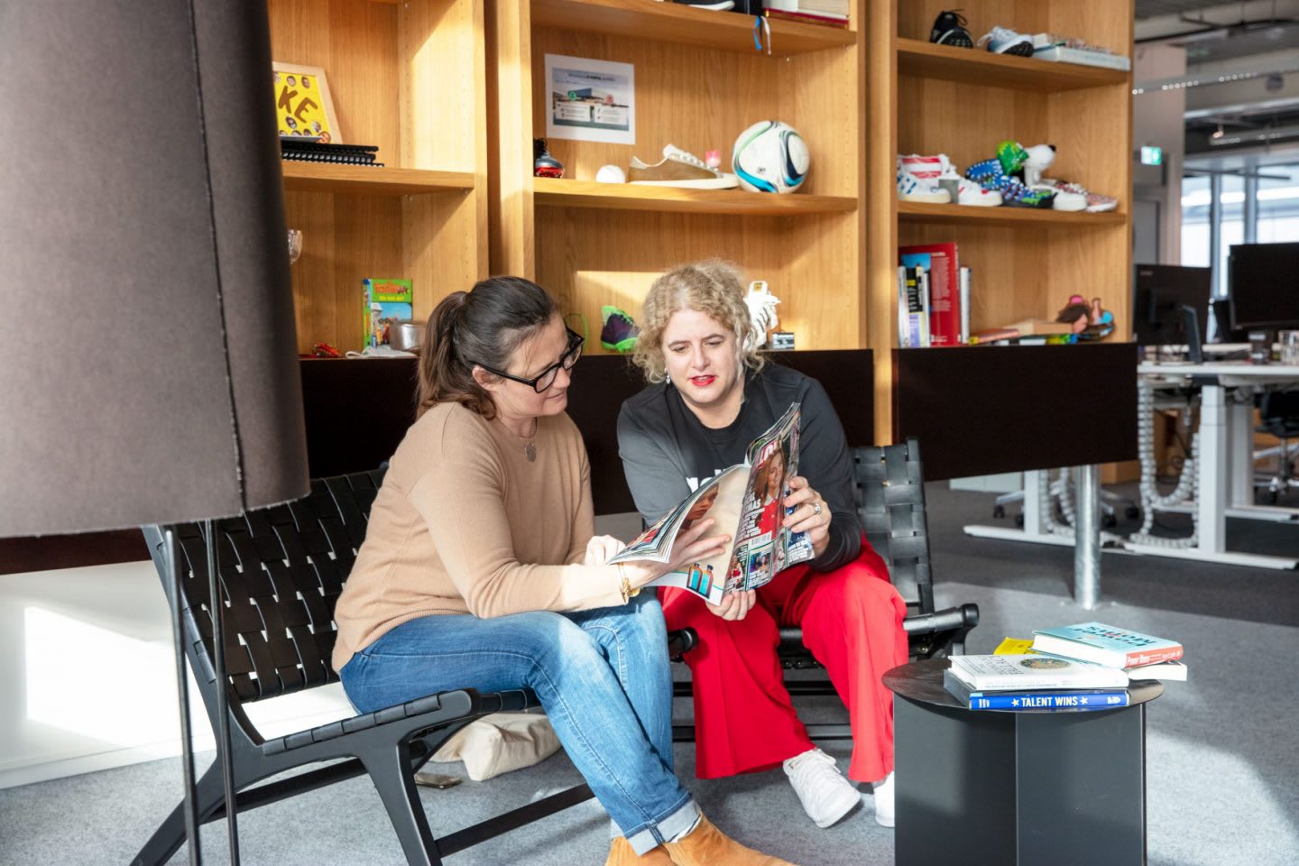 Two women sit on a bench in an office environment reading together.