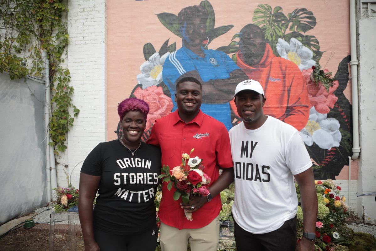 Three people stand in front of a vibrant wall mural celebrating Black excellence.