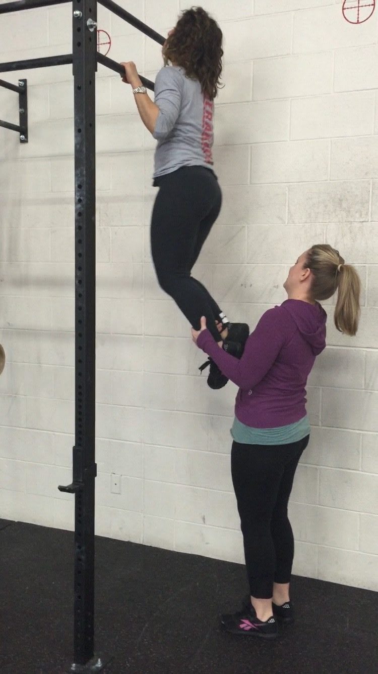 Two-Women-Helping-Each-Other-While-Doing-Pullups-White-Wall-Indoors-Gym
