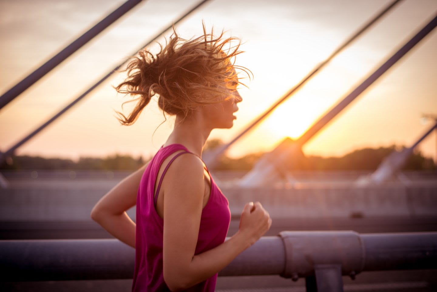 Young sportswoman jogging over the bridge in the morning while her long brown hair is waving in the wind. Rising sun is in the background.