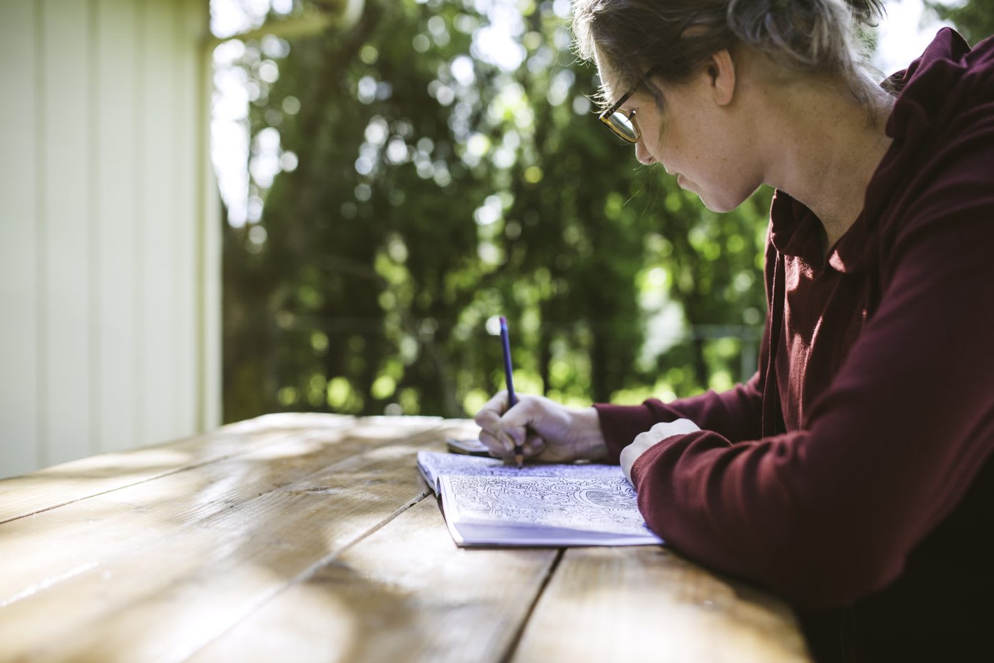 A woman enjoys the relaxation and calming benefits of using a coloring book, selecting different colored pencils to fill in an intricate design sitting at a wooden table. doodling, relaxing, passing time, writing, habit, hobbies.