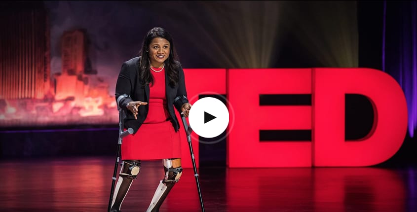 Woman in red dress with prosthetic legs and crutches giving speech on stage in front of red sign, TED Talks, challenges, inspirational, speaker, talk, GamePlan A