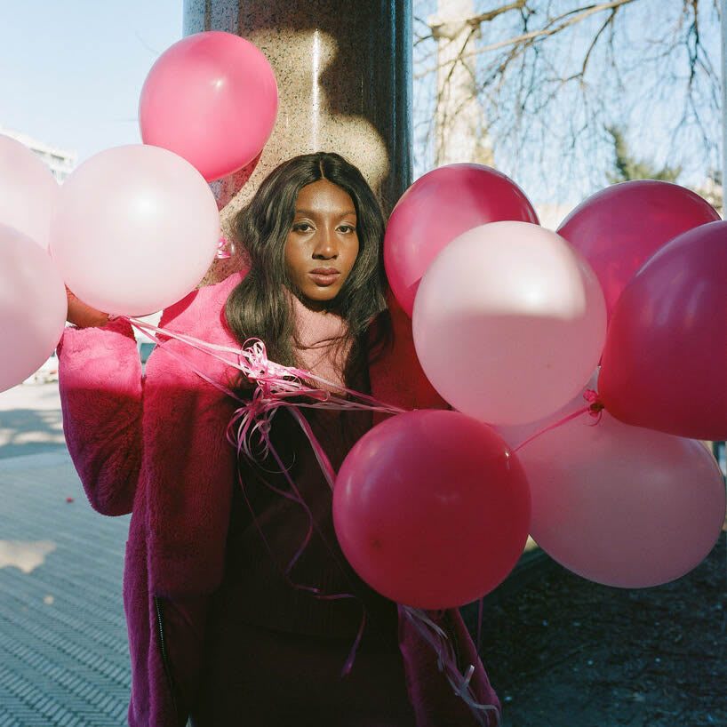 Woman holding pink and red balloons posing and staring at the camera, race, Ziwe May, inclusion, podcast