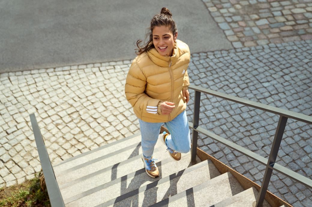 Woman wearing beige adidas puffer jacket and jeans running up stairs, woman, adidas, athletic, lifestyle, activewear, sports, sport