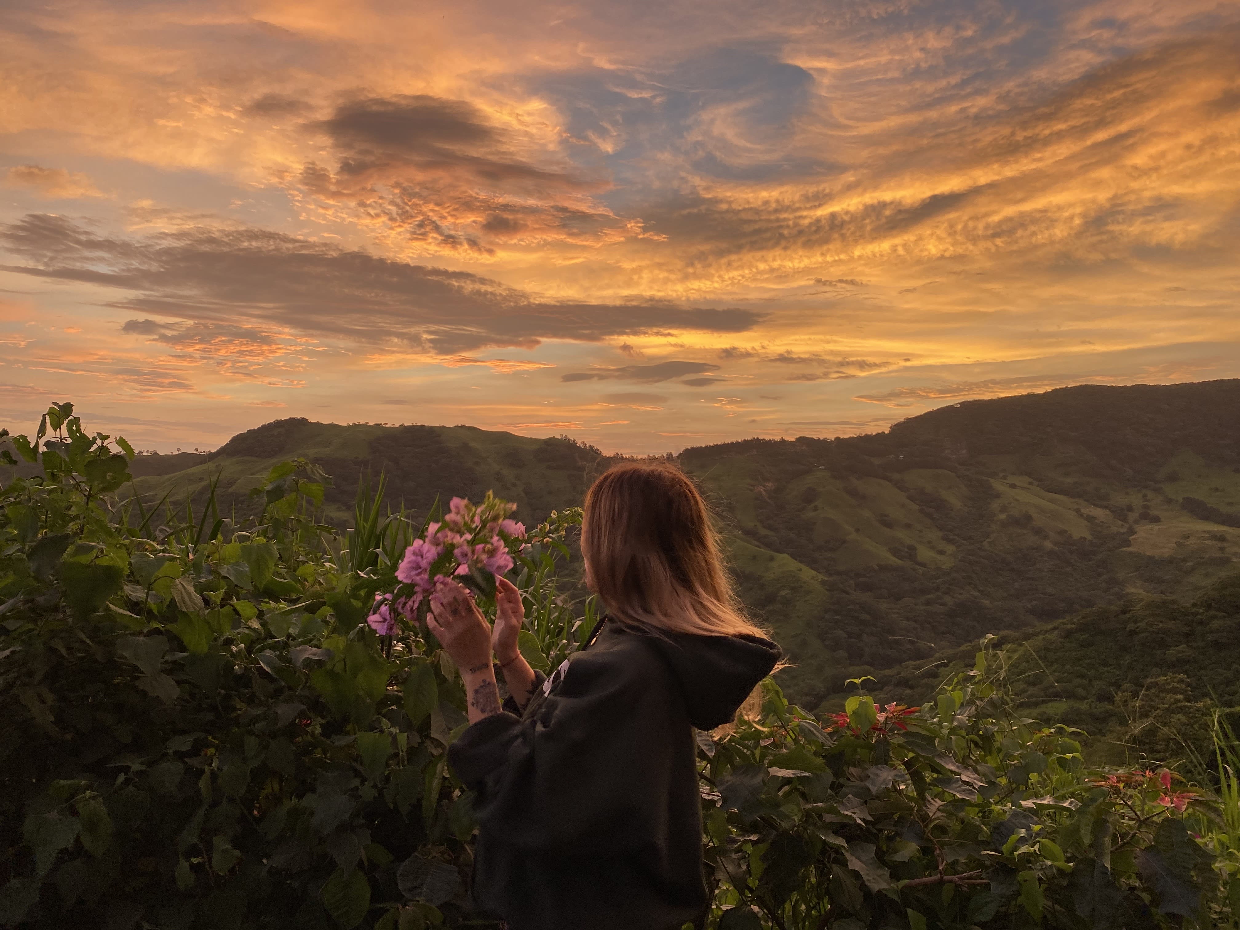 Luz holding wildflowers while gazing at a vibrant mountain sunset