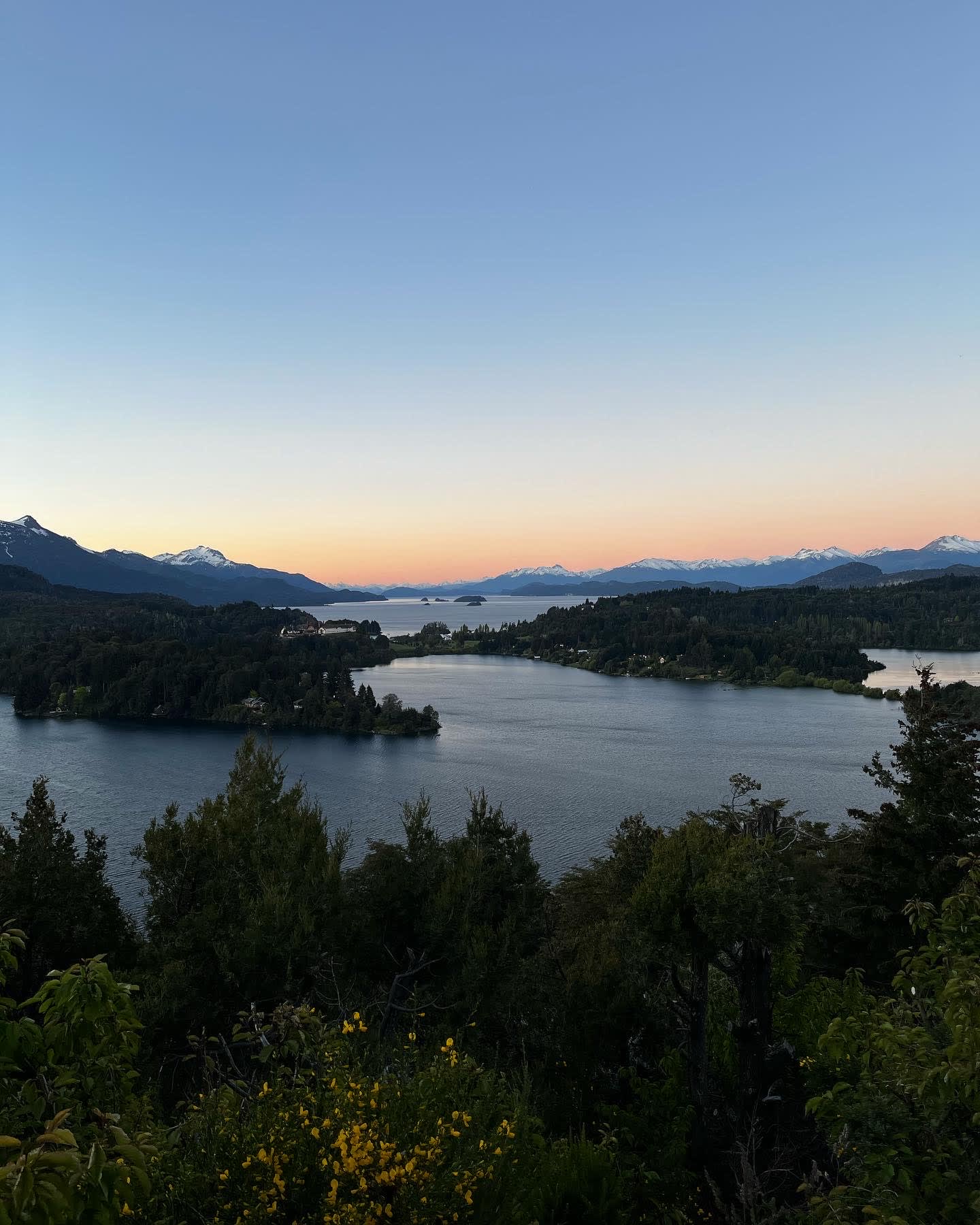 Panoramic mountain view overlooking large lakes surrounded by forests and snow-capped peaks beneath a sunset sky