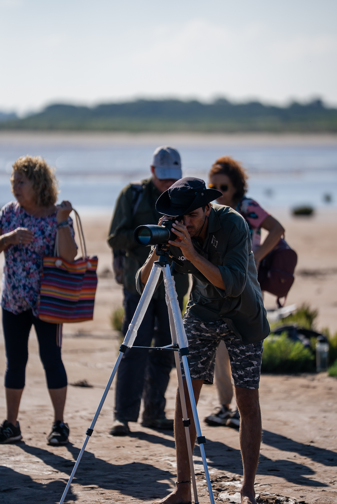 Biólogo observando aves con un monocular junto a turistas y un río detrás.