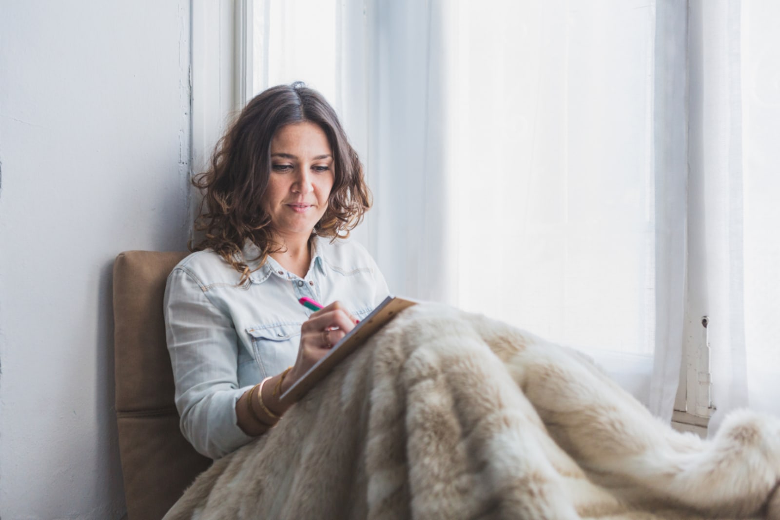 A woman writing in a journal by a bright window, wrapped in a soft blanket