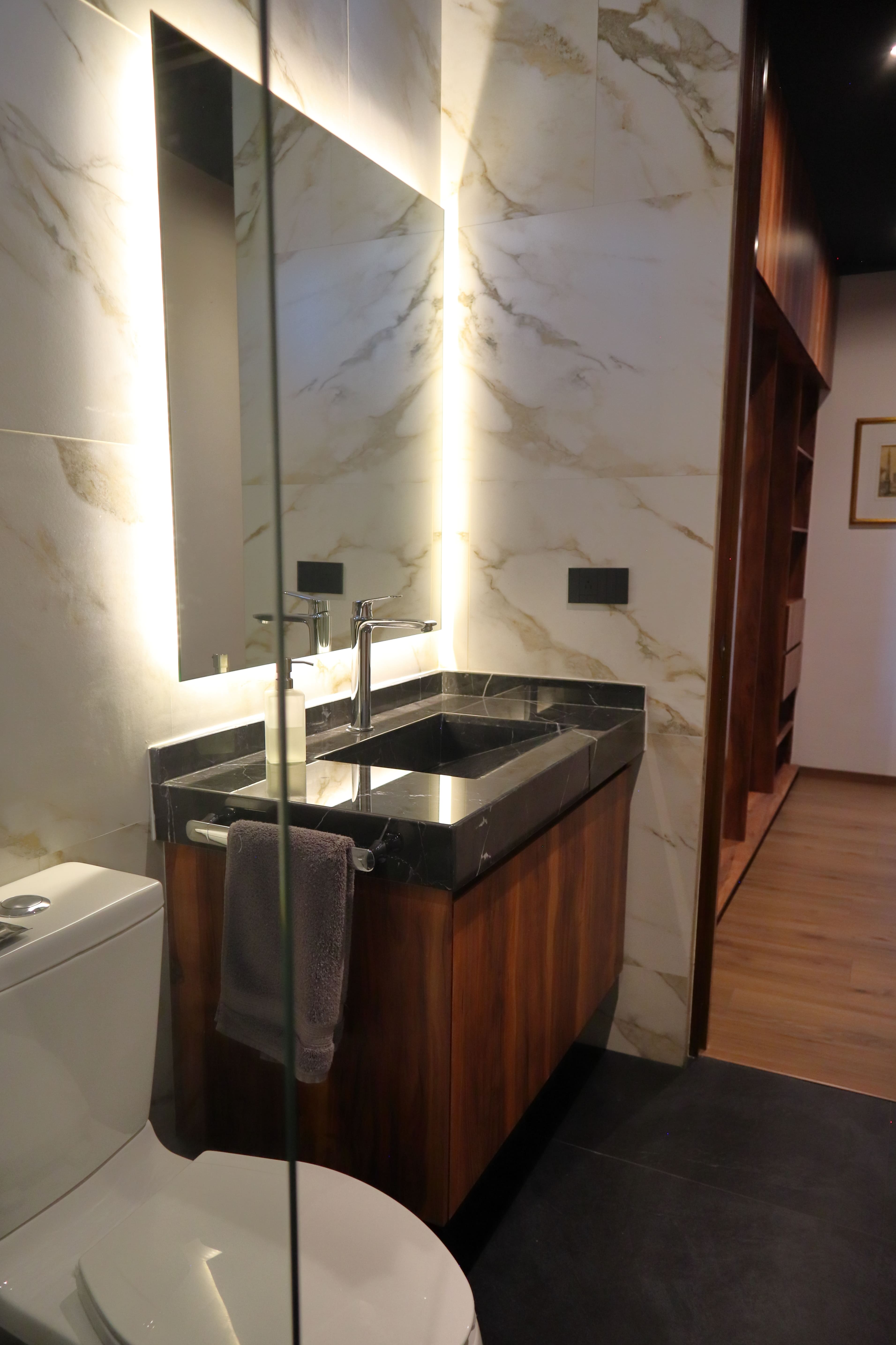 Diseño de baño para recámara secundaria en Casa DRM, mostrando un lavabo de granito oscuro sobre un mueble de madera y un muro de mármol blanco con iluminación LED vertical tras el espejo. Proyecto de MGDC en Campestre, SLP.