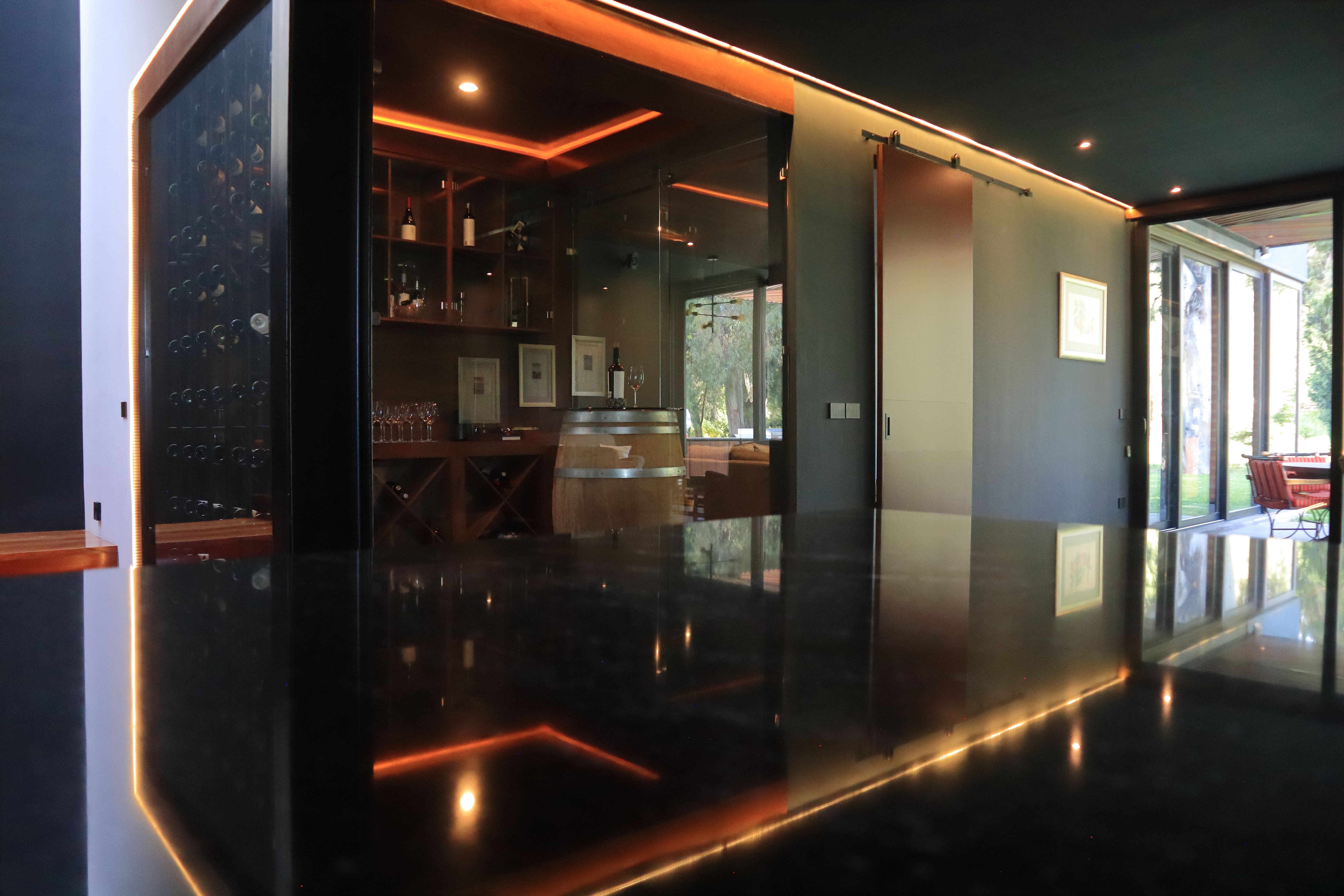 Interior de la Casa DRM mostrando la cava de vinos de lujo con iluminación cálida, reflejada en el piso negro brillante, y grandes ventanales de cristal que conectan el espacio con la terraza exterior. Proyecto de MGDC en Campestre, SLP.