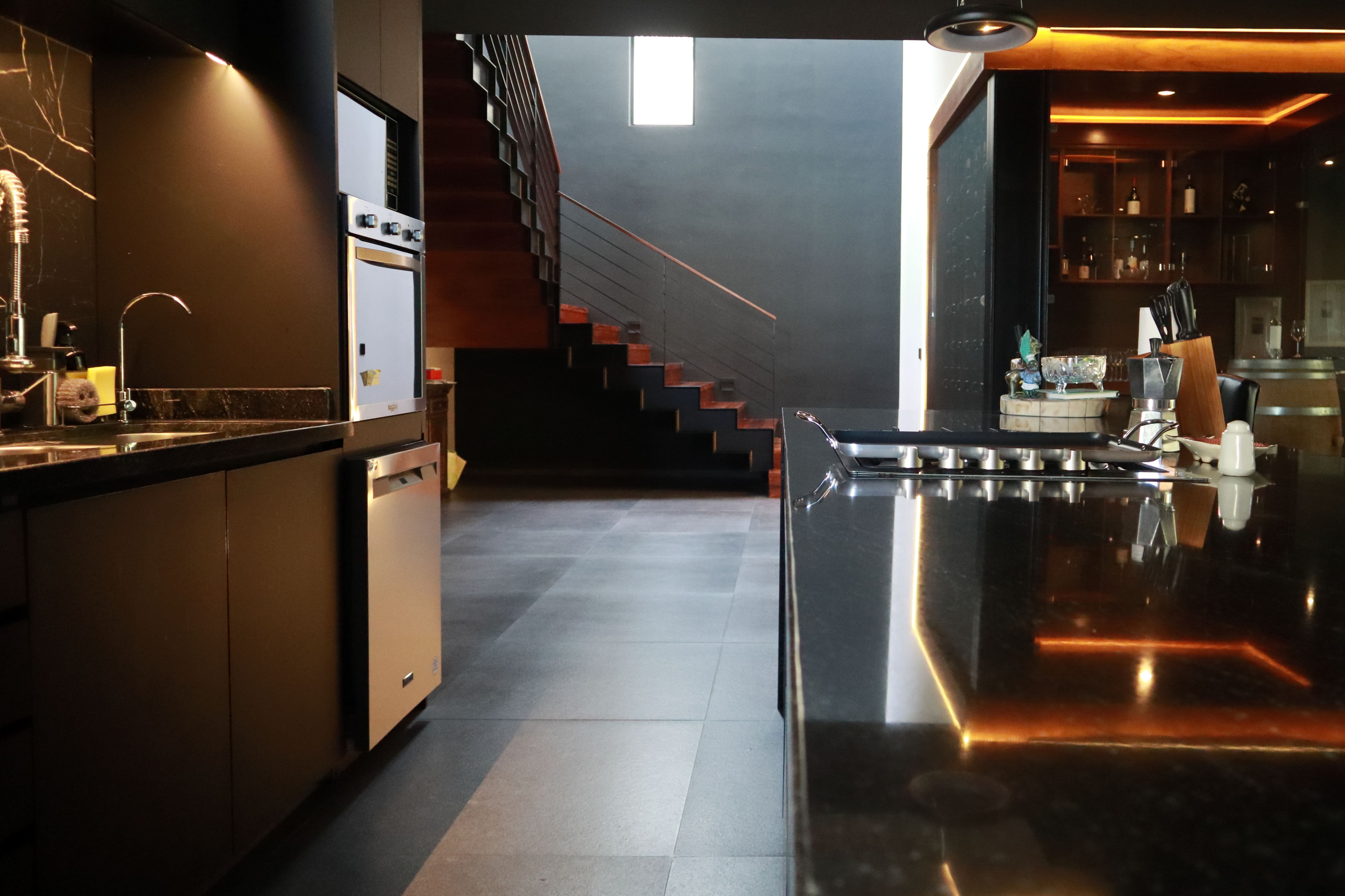 Vista interior de la cocina de lujo en Casa DRM, mostrando la cubierta de granito negro brillante, con las escaleras y la cava al fondo. Proyecto residencial de MGDC en Campestre, SLP.