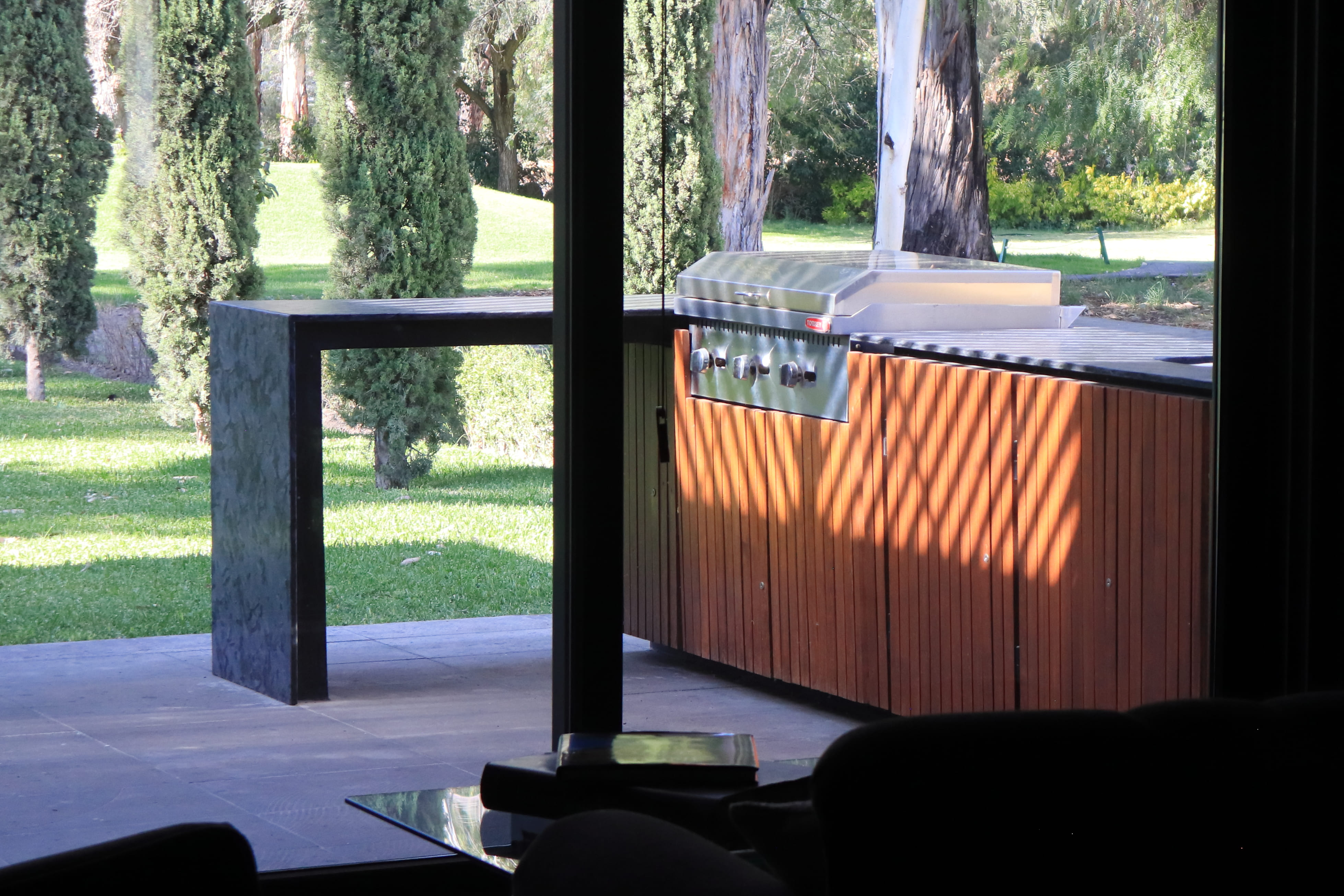 Vista desde el interior de la casa hacia la terraza con asador de acero inoxidable empotrado en un mueble de madera y una barra de concreto, con el jardín de fondo. Proyecto Casa DRM por MGDC en Campestre, SLP.
