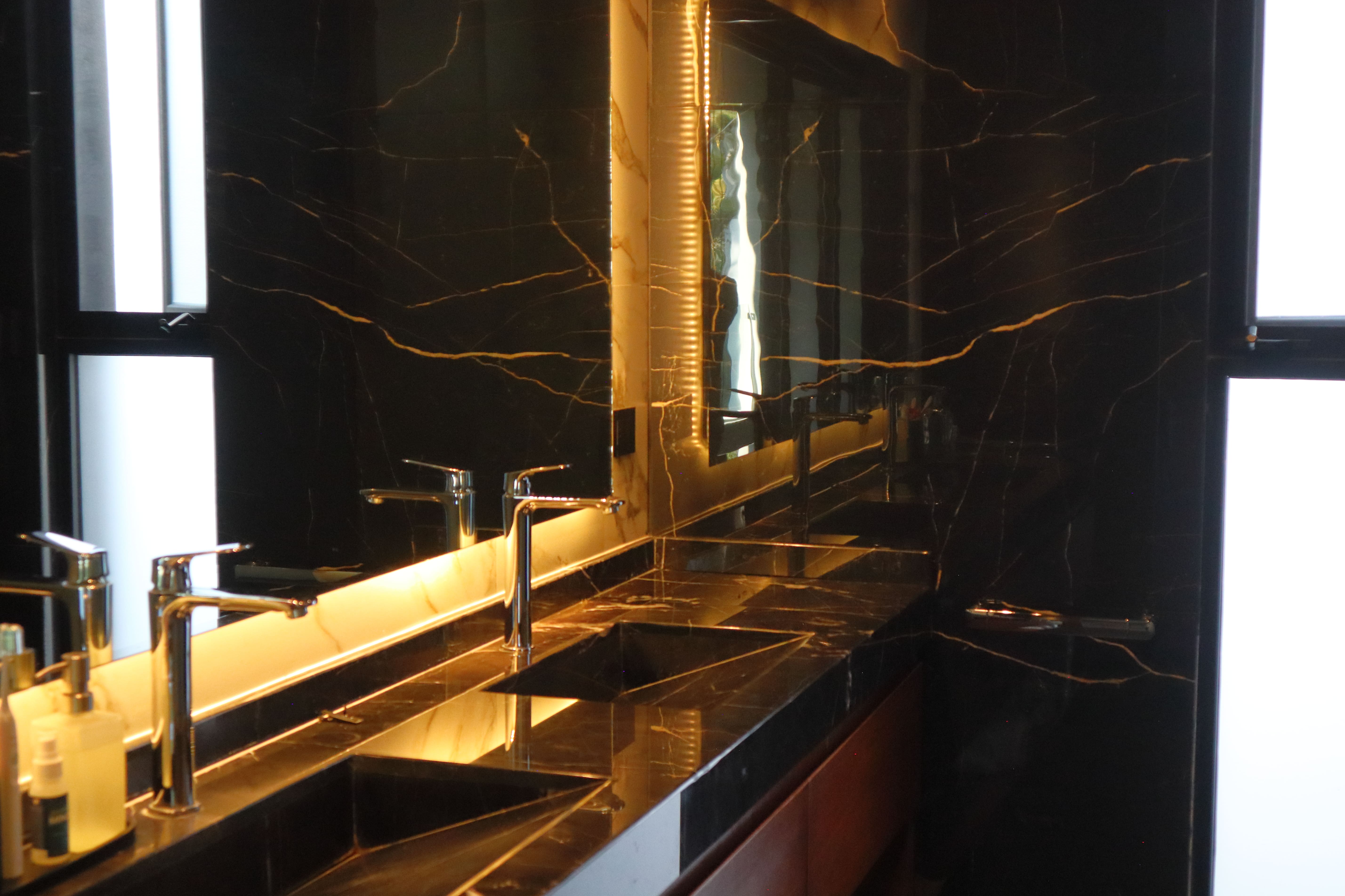 Diseño del baño de la recámara principal de Casa DRM, mostrando un lavabo doble sobre una cubierta de mármol negro veteado que continúa en los muros. La iluminación indirecta del espejo resalta los acabados de lujo. Proyecto de MGDC en Campestre, SLP.