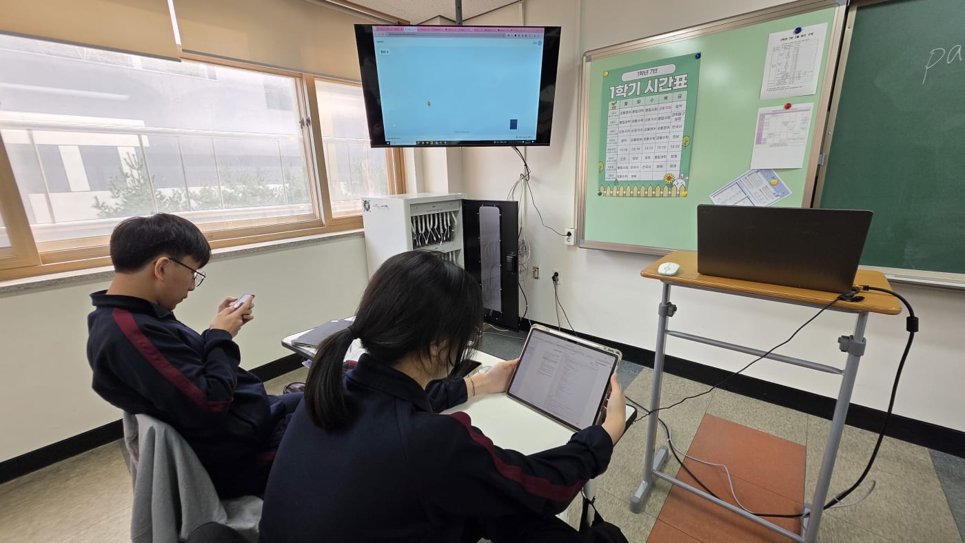 교실 전면에 수업 자료가 띄워져 있으며 학생들이 개인 기기로 코딩 오류를 해결하며 논리적 사고를 검증하는 장면입니다.