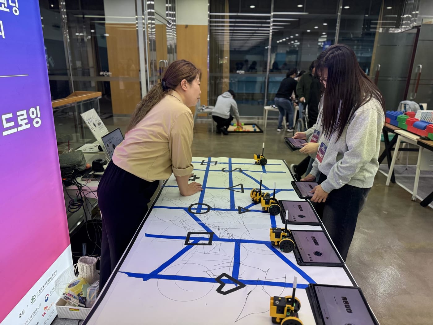 탁상 위의 대형 맵 위에서 소형 로봇과 태블릿 PC를 활용해 실전 코딩을 수행하는 학생과 교육자의 모습입니다. 기술적 호기심과 실습의 역동성이 느껴지는 현장입니다.