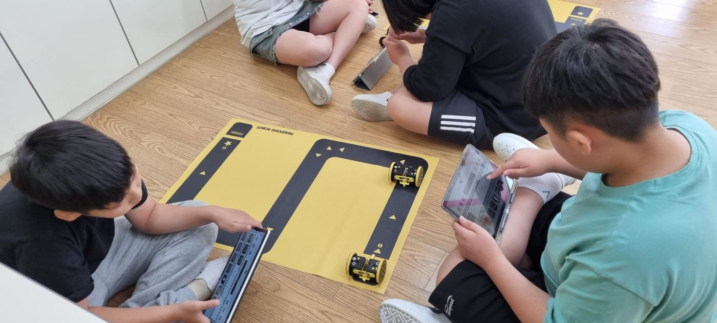 바닥에 앉아 교보재와 태블릿을 이용해 로봇의 경로를 코딩하는 어린이들의 모습입니다. 자유로운 분위기 속에서 즐겁게 학습하는 모습이 인상적입니다.