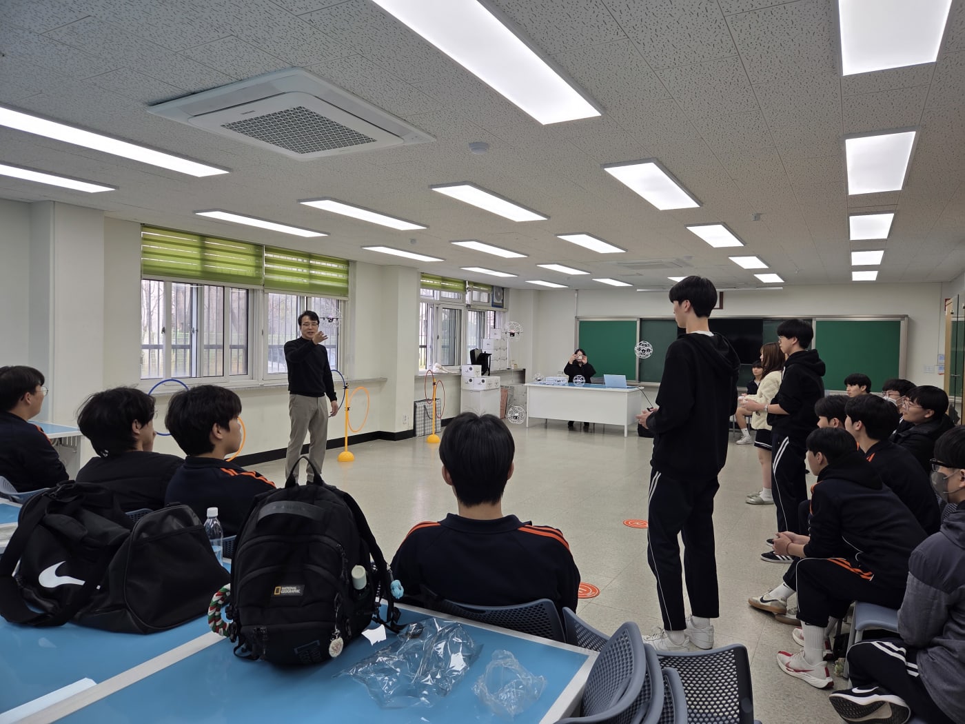 강사가 학생들 앞에서 드론 비행을 직접 시연하며 실습 요령을 설명하는 장면입니다. 교실 내부에 비행 장애물인 후프가 설치되어 있어 실습의 역동성이 느껴집니다.