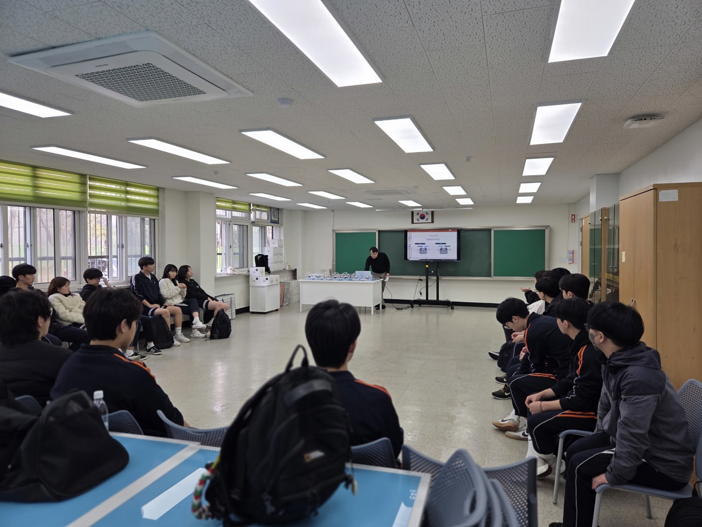 강의 자료를 점검하며 수업을 준비하는 강사와 이를 경청하기 위해 대기하는 학생들의 모습입니다. 전반적으로 깔끔하고 정돈된 교실 분위기가 안정감을 줍니다.