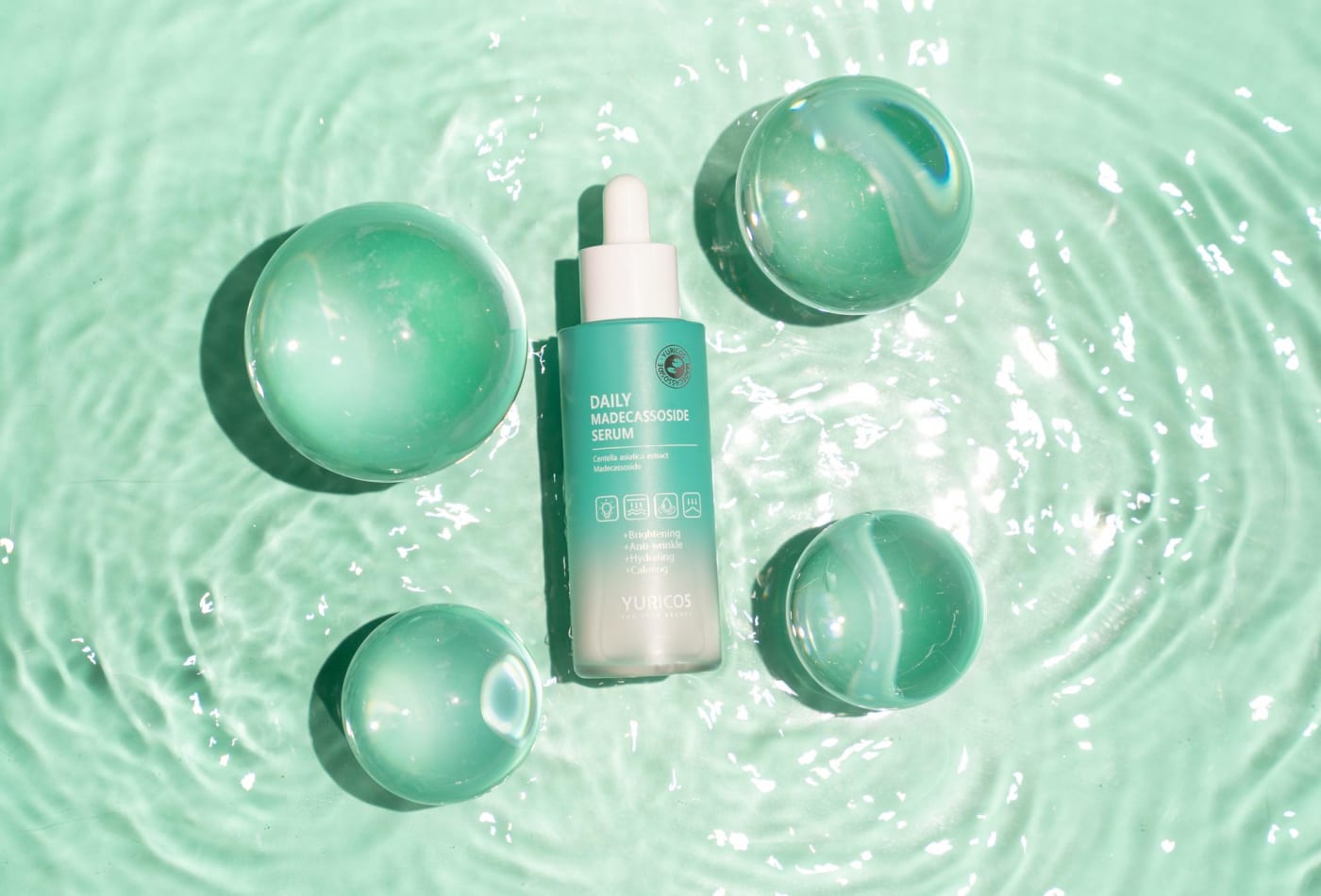 The daily Madecassoside Serum in a refreshing mint color package is placed amidst clear water ripples and glass beads, conveying its powerful moisturizing and soothing effects visually.