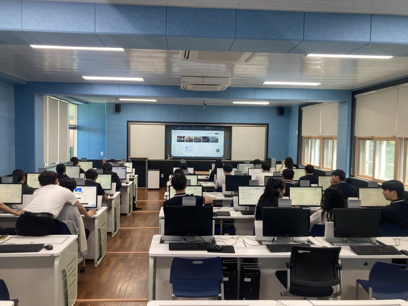 강의실 전면의 대형 스크린에 AI 기반 창작 도구가 띄워져 있으며, 학생들이 일제히 집중하고 있는 체계적인 학습 전경입니다.