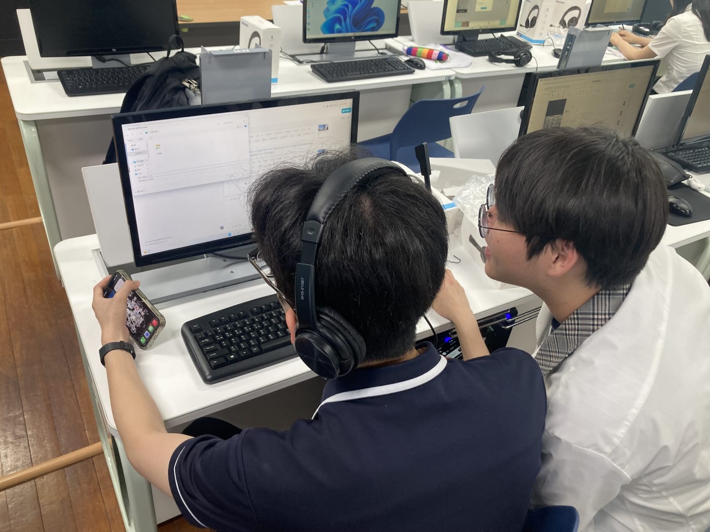 두 학생이 머리를 맞대고 모바일 기기와 PC 화면을 번갈아 보며 제작 중인 영상 결과물에 대해 소통하는 협력 실습 장면입니다.
