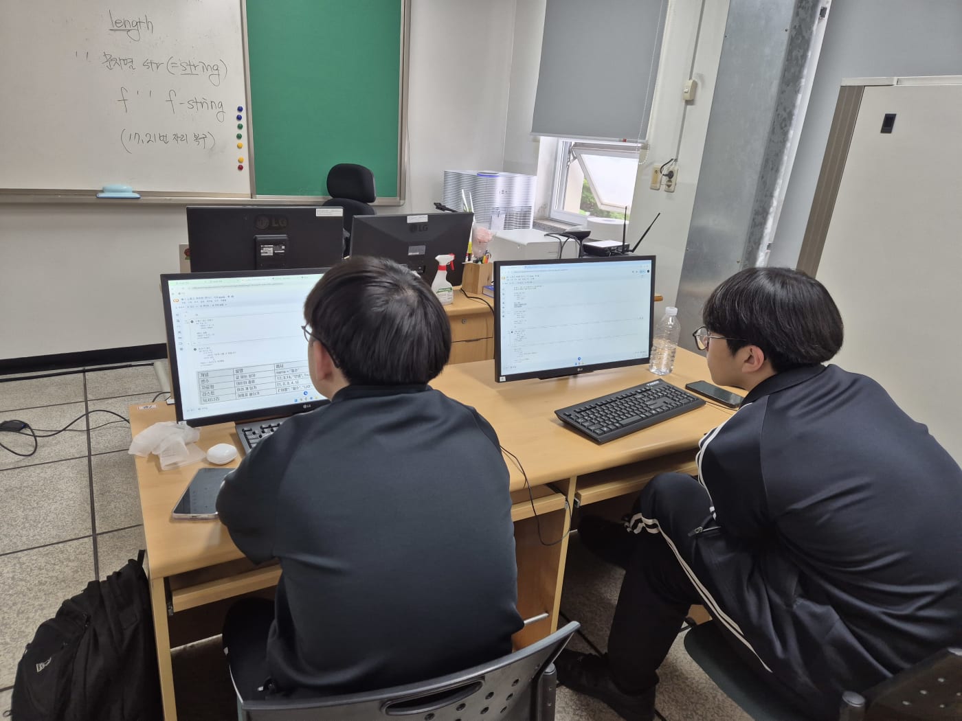 화이트보드에 적힌 f-string 문법 설명과 모니터상의 코드를 대조하며 심도 있게 공부하는 학생들의 모습입니다.