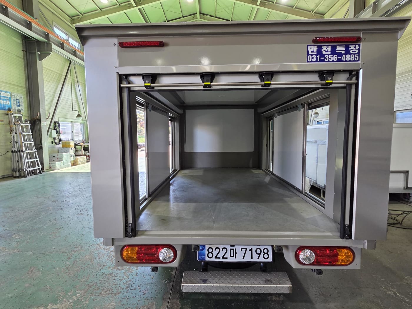 후면에서 바라본 탑차의 개방된 모습입니다. 상단 셔터 도어가 부드럽게 열려 넓은 적재 입구를 확보하고 있으며, 차량 하부의 견고한 설계와 내부의 청결한 상태가 잘 드러납니다.