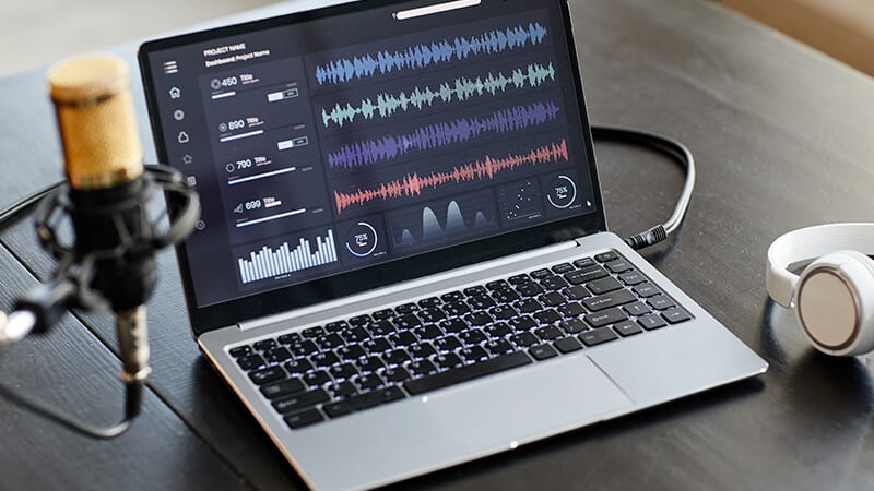 Imagem de um microfone focado e ao fundo uma tela de notebook com ondas de áudio.