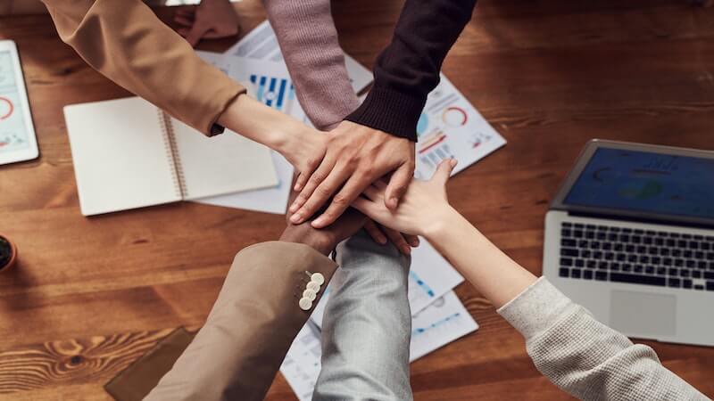 The image shows six people with hands together, above a desk, in a sign of teamwork