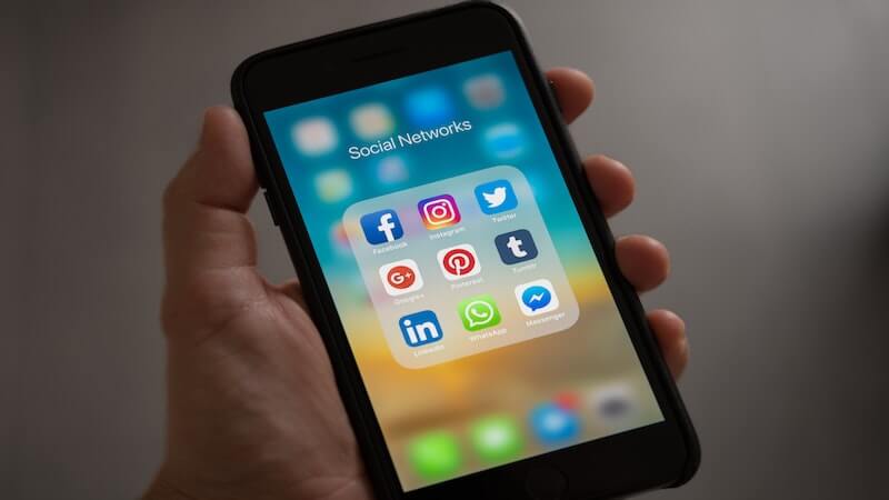 The image shows a person holding a smartphone, on the screen there are several social media icons, such as Facebook, Instagram, Twitter, and LinkedIn.