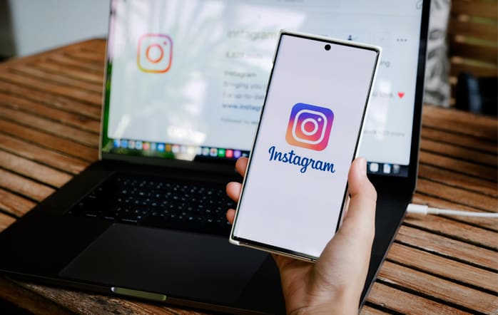Foto de uma pessoa segurando um celular com Instagram aberto e um notebook ao fundo