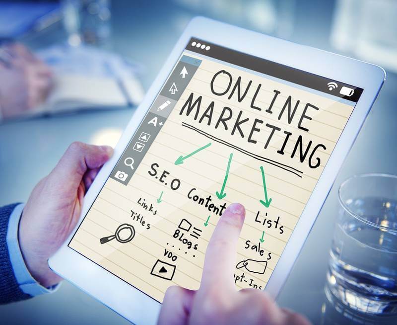 In the image there is a person holding a tablet, and on its screen it's written "Online Marketing", with arrows pointing to the words "SEO", "Content", and "Lists".