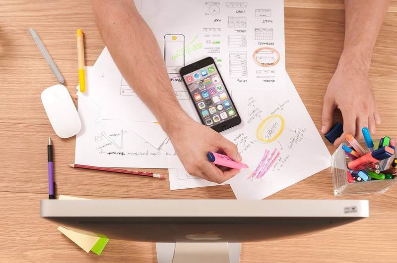 The image shows a person highlighting a paper on a table, in front of the person there is a desktop computer. On the table there are a lot of papers with diagrams, a smartphone and pens. 