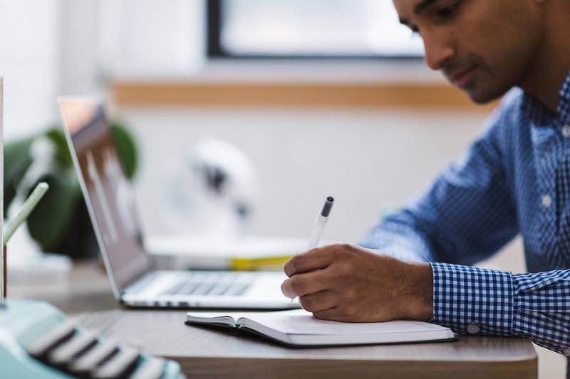 The image shows as man sitting on a table in front of a computer. He is majing notes on a notebook.