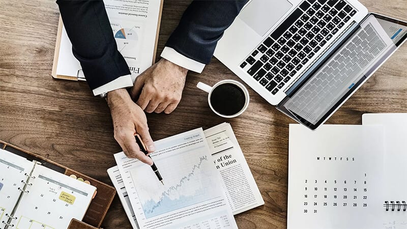 Foto de uma pessoa em analisando gráficos em cima de uma mesa com notebook e café