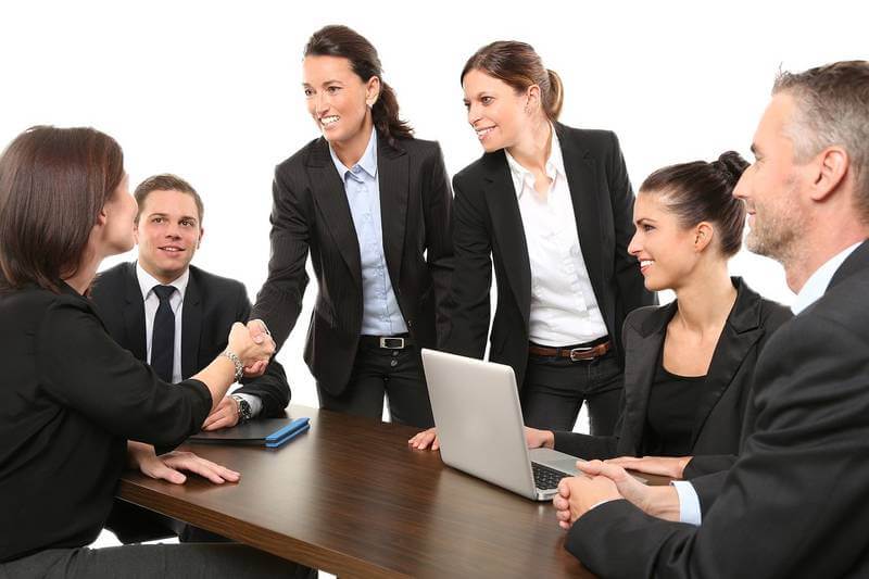 The image shows a group o teamworks sitting around a table, two women are standing and one of them shake the hand of another woman sitting in front of her.