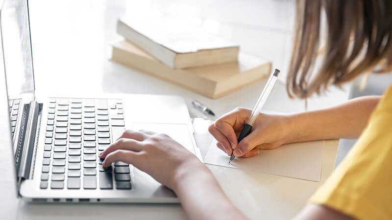 Foto de uma mulher fazendo anotações em um caderno e utilizando um notebook