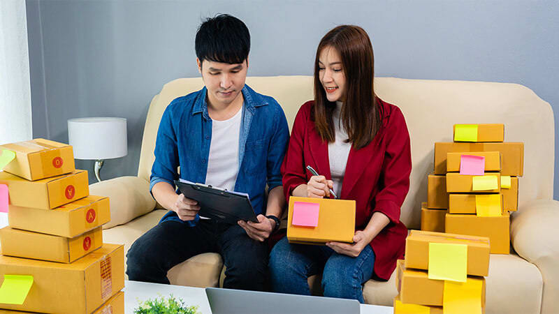 The image shows a man and a woman sitting on a couch. The man is holding a tablet and the woman is holding a box. On the side of the couch and on the table in front of them there are several boxes.