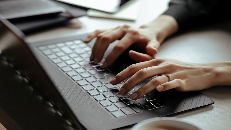 Foto de um notebook em cima da mesa e duas mãos femininas usando as teclas