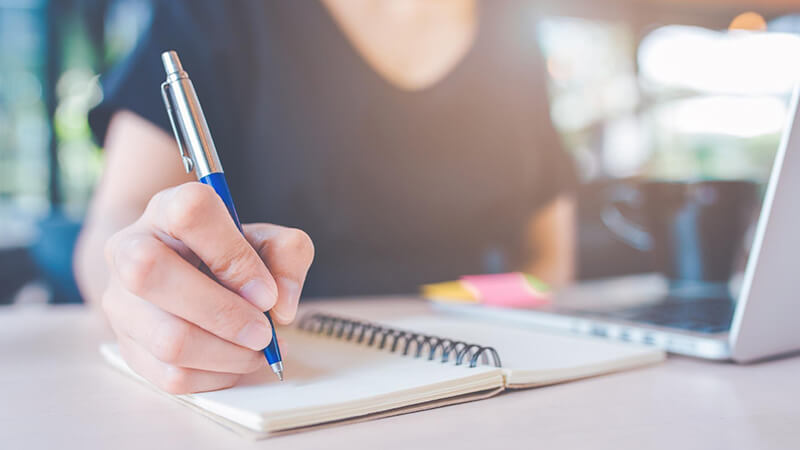 Foto de uma mulher utilizando um notebook e fazendo anotações em um caderno