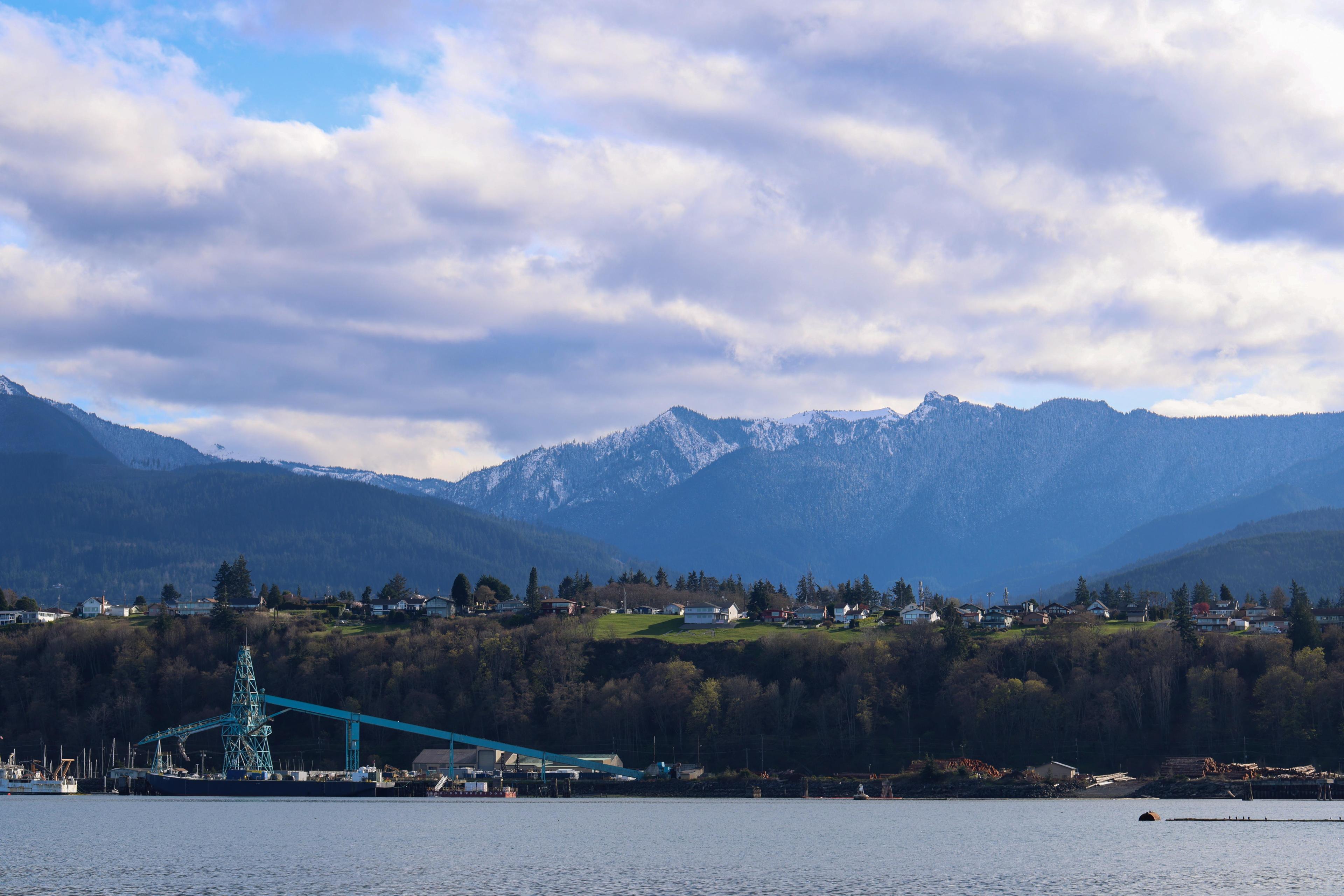 Snow-capped Olympic Mountains rising behind Port Angeles harbor with industrial waterfront and cloudy spring sky