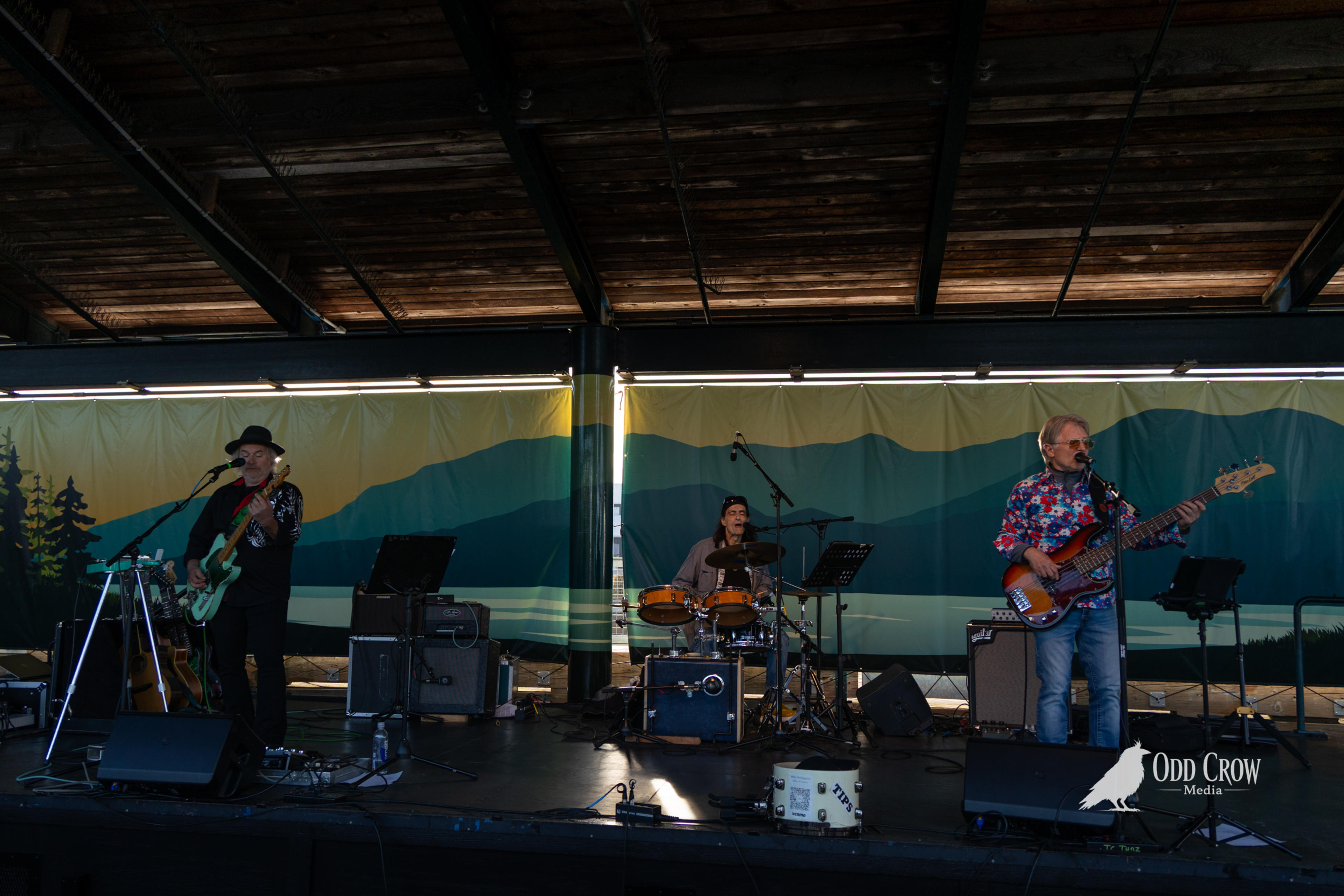 Three-piece band The Foot Stompers performing on stage at Concerts on the Pier in Port Angeles, Washington, with a painted Pacific Northwest mountain backdrop behind them