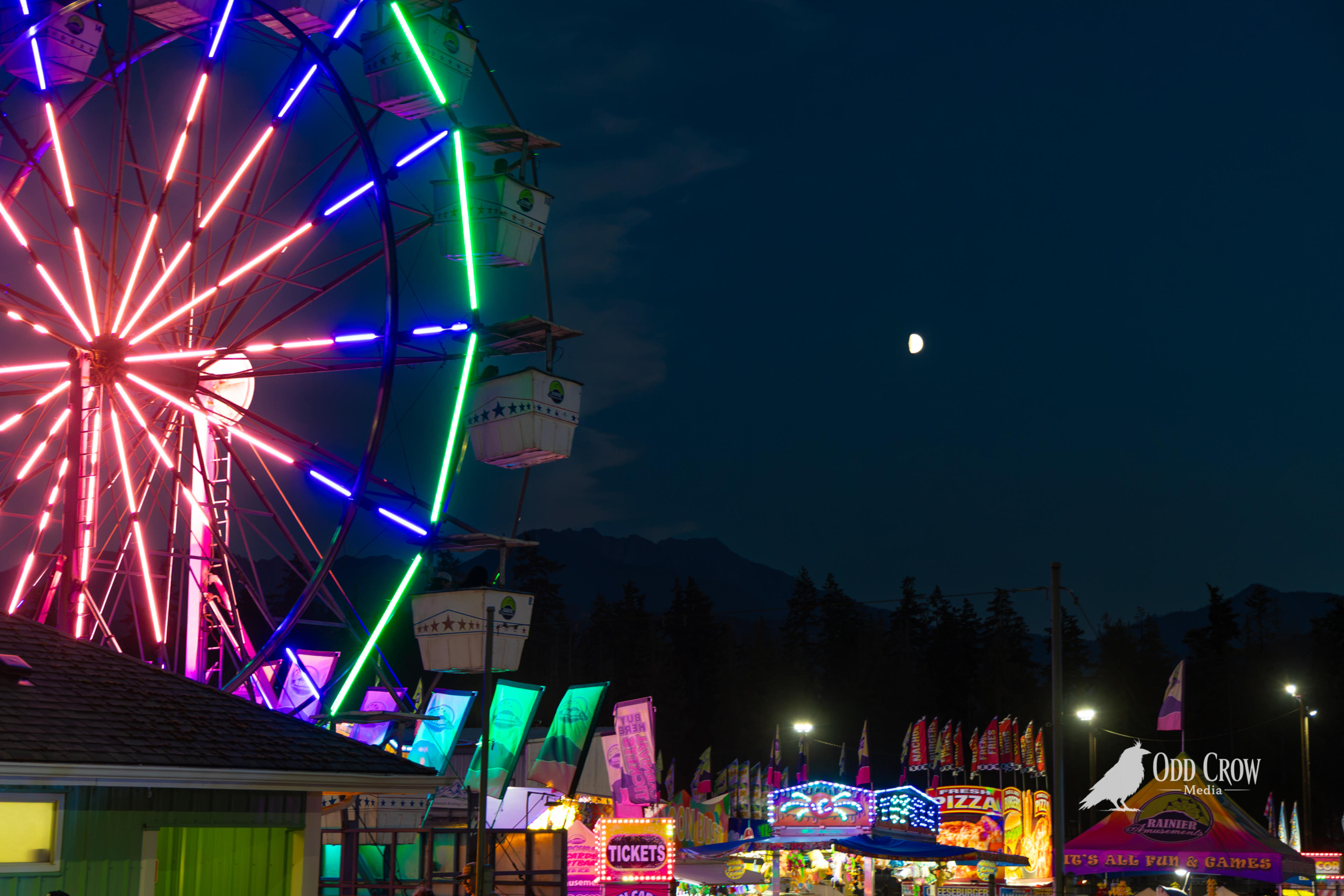 Clallam County Fair Ferris wheel lit up in neon colors at night with a half moon visible in the dark blue sky above the Olympic Mountains
