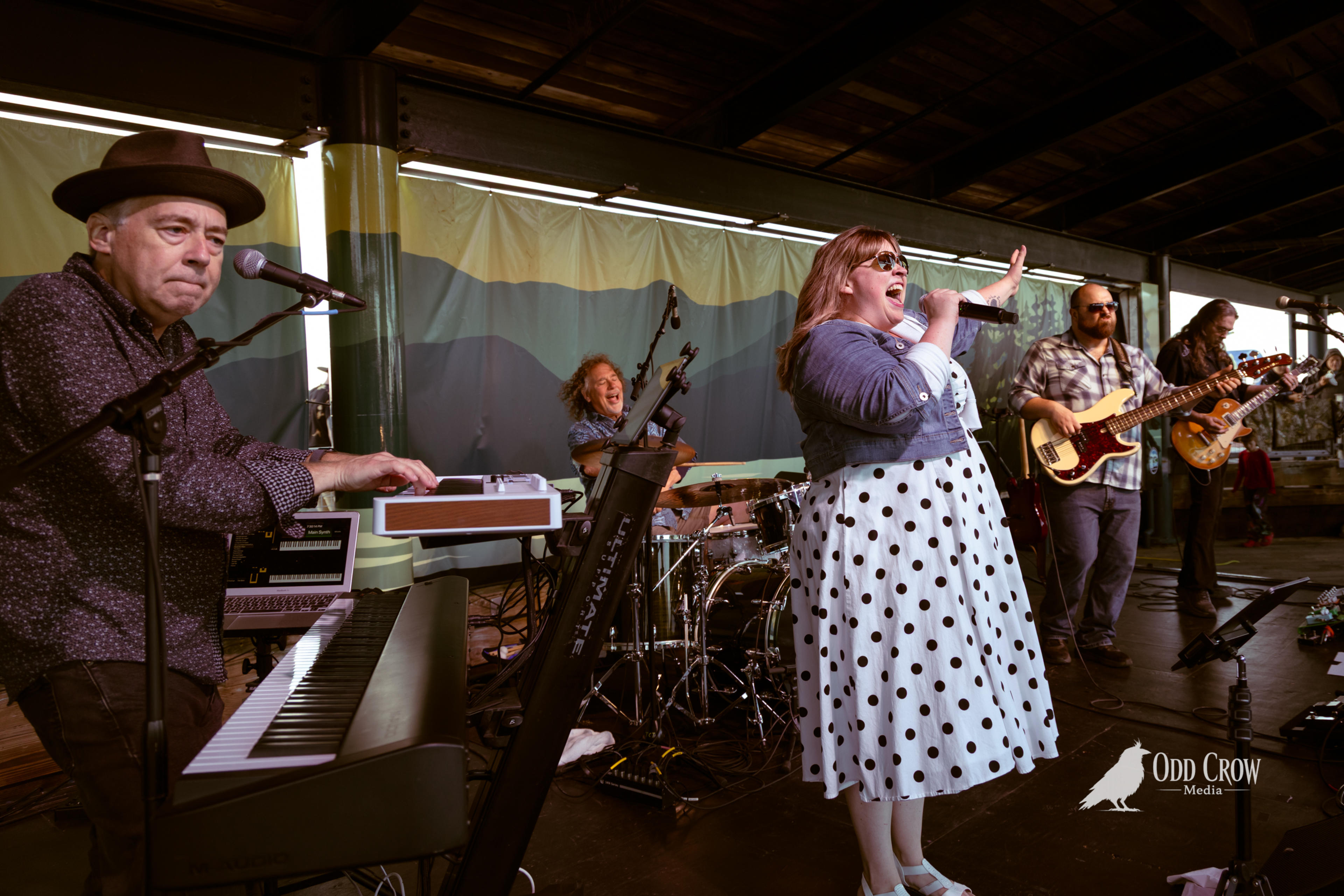Female vocalist in white polka dot dress performing on stage at Concerts at the Pier, Port Angeles, with crowd dancing in background at golden hour